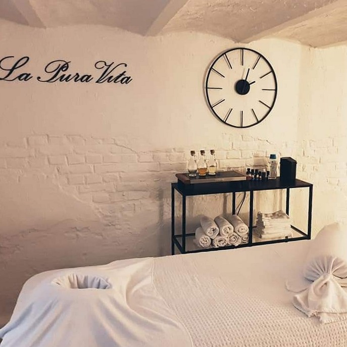 De behandelruimte met massagetafel, opgerolde handdoeken, een klok en de naam La Pura Vita op de muur